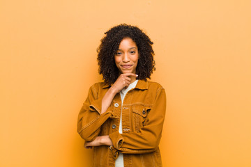 young pretty black woman looking happy and smiling with hand on chin, wondering or asking a...
