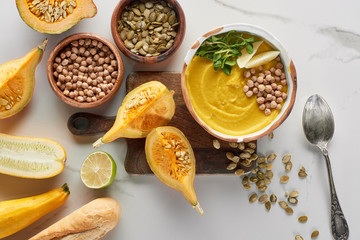 top view of autumnal mashed pumpkin soup in bowl on wooden cutting board near ingredients on marble surface