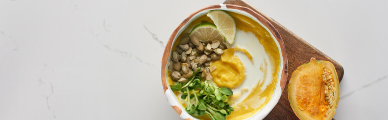 top view of autumnal mashed pumpkin soup in bowl on wooden cutting board with pumpkin on marble surface, panoramic shot