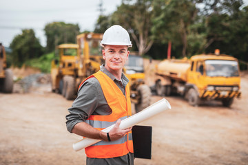 Caucasian handsome engineer is stand and smile holding a laptop with roll of paper on road...