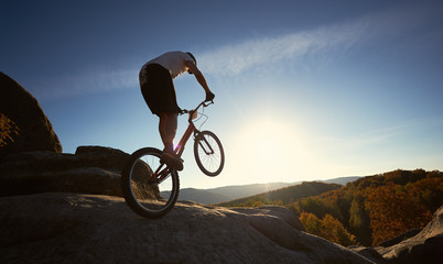 Silhouette of professional sportsman cyclist riding on back wheel on trial bicycle, man rider making acrobatic trick on the big boulder in the summer sunny evening. Blue sky and sunset on background.