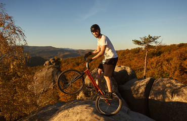 Fototapeta premium Professional sportsman cyclist riding on back wheel on trial bike. Male athlete rider looking to the camera, making acrobatic stunt the top of mountain. Forest and evening blue sky on background