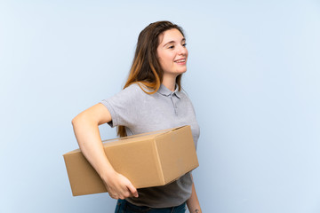 Young brunette girl over isolated blue background holding a box to move it to another site in...