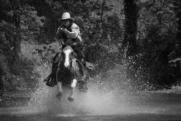 A vintage action shot of a cowboy riding a horse, wading through the water and holding a rifle.