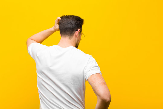 young handsome man feeling clueless and confused, thinking a solution, with hand on hip and other on head, rear view against orange wall