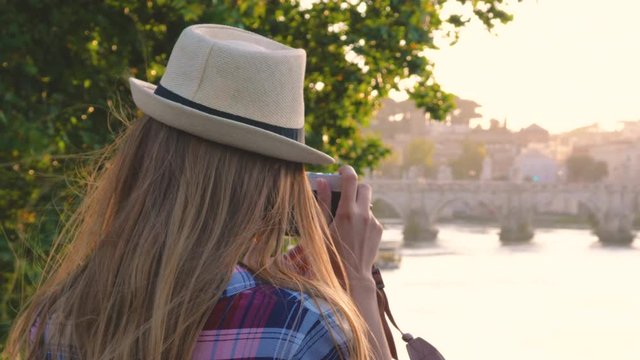 Young blonde woman tourist with hat on a bridge on river tiber in Rome, taking pictures with vintage camera of St. Peter and Vatican city, Italy.