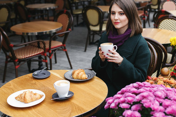 Beautiful young girl wearing green coat sitting at a table in cozy street outdoor cafe and drinking coffee with a croissant. Restaurant terrace is decorated with chrysanthemum flowers bushes in autumn