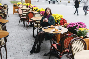 Beautiful young girl wearing green coat sitting at a table in cozy street outdoor cafe and drinking...