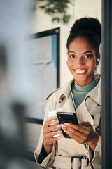 Fototapeta premium Attractive casual African American girl in stylish trench coat with cellphone joyfully looking in camera at bus stop