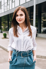 attractive young woman in white shirt standing with hands in pockets on street