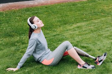 disabled sportswoman sitting on grass and listening music in headphones