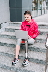 smiling disabled sportswoman using laptop while sitting on stairs on street
