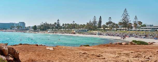 Fototapeta na wymiar Panoramic shot of a beach with stones and transparent water of the azure mediterranean sea, surrounded by a picturesque nature of Cyprus. Ayia Napa.