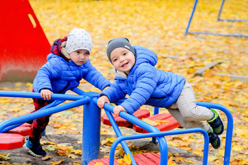 Brightly dressed twin brothers play on a carousel in an autumn park amid fallen yellow leaves. They are two years old.