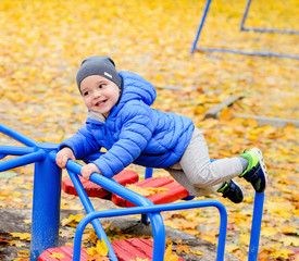 A brightly dressed two-year-old boy is playing on a carousel in an autumn park. Bright blue carousel and yellow fallen leaves.