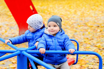 Brightly dressed twin brothers play on a carousel in an autumn park amid fallen yellow leaves. They are two years old.