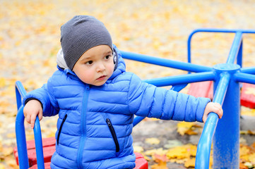 A brightly dressed two-year-old boy is playing on a carousel in an autumn park. Bright blue carousel and yellow fallen leaves.
