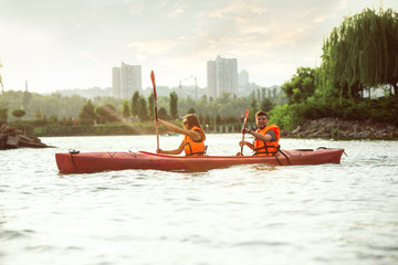 Happy young caucasian couple kayaking on river with sunset in the backgrounds. Having fun in leisure activity. Happy male and female model laughting on the kayak. Sport, relations concept. Colorful.