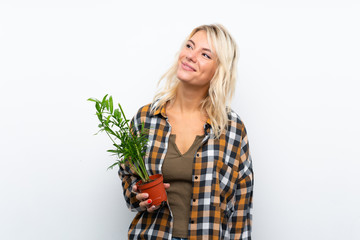 Young blonde gardener woman holding a plant over isolated white background looking up while smiling