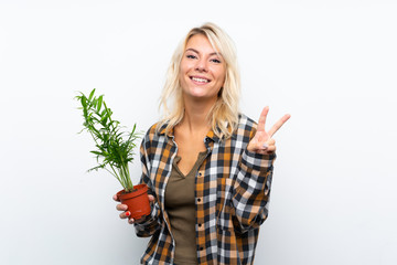 Young blonde gardener woman holding a plant over isolated white background smiling and showing victory sign