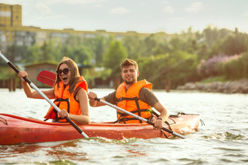 Happy young caucasian couple kayaking on river with sunset in the backgrounds. Having fun in leisure activity. Happy male and female model laughting on the kayak. Sport, relations concept. Colorful.