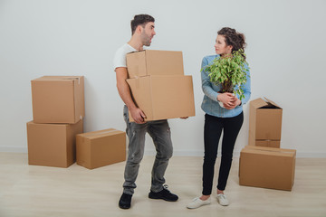 Man can not hold heavy carton boxes anymore, but he doesn't know where to put them and is shouting at his wife. His wife is standing with flowerpot in hands, happy face expression.