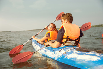 Fototapeta na wymiar Happy young caucasian couple kayaking on river with sunset in the backgrounds. Having fun in leisure activity. Happy male and female model laughting on the kayak. Sport, relations concept. Colorful.