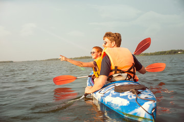 Happy young caucasian couple kayaking on river with sunset in the backgrounds. Having fun in leisure activity. Happy male and female model laughting on the kayak. Sport, relations concept. Colorful.