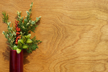 Horizontal flat lay of a slender red vase with colorful evergreens and bright red winterberry holly berries on polished oak wood background, with copy space