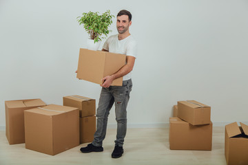 Handsome young man moving into new house, standing, holding carton box and flowerpot, smiling, surrounded by boxes.