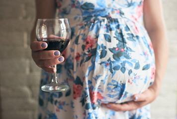 Selective focus of glass of wine in hands of pregnant woman. Future mother embracing belly and...