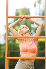 Fototapeta premium Sexy young fitness girl pulls up on street sport gym. Blonde fitness woman in sport wear with perfect fitness body on street workout performing abdominal exercises on the horizontal bar