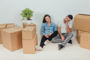 Fototapeta na wymiar The process of moving into new house is totally exhausting. Lovely young couple wife and husband are sitting on the floor in new apartment among carton boxes.