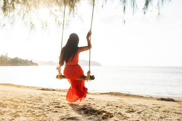 girl in a red dress on a swing among tropical palm trees in Thailand at sunset