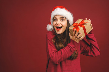Excited surprised woman in red santa claus outfit holding present isolated on the red background.