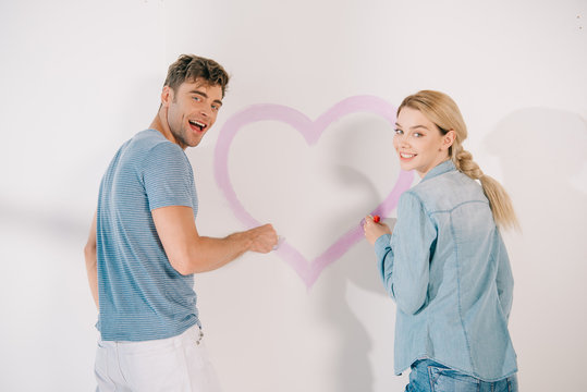 happy young couple smiling at camera while standing near pink heart drawn on white wall