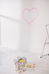 room with pink heart drawn on white wall, cans with paint and paintbrushes on white floor covered with cellophane