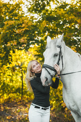 Young smiling blond woman hugs a white horse in a autumn forest.