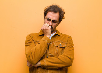 young handsome man feeling serious, thoughtful and concerned, staring sideways with hand pressed against chin against orange wall
