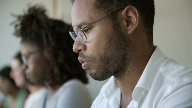 Focused African American man in eyeglasses looking down. Concentrated colleagues sitting in row and working. Employment concept