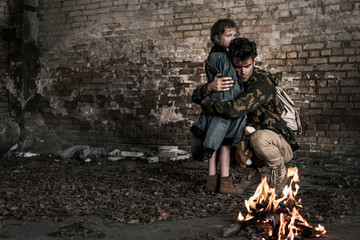 handsome man hugging child near burning bonfire, post apocalyptic concept