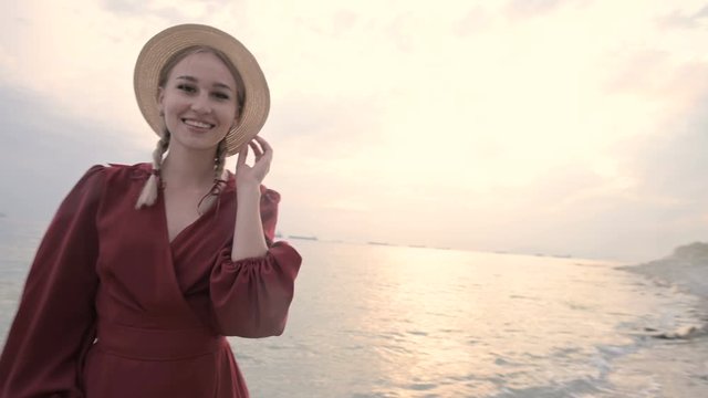 Young attractive smiling girl in a red light dress and a straw hat whirls walking along the seashore in windy weather at sunset. Femininity and independence in lightness and beauty