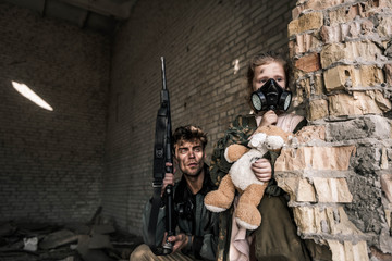 kid in gas mask holding teddy bear near handsome man with gun, post apocalyptic concept