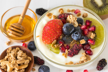 Plain yogurt with strawberry, blueberries, kiwi, granola, pomegranate in a glass bowl and honey on white wooden texture, healthy food and plant-based food concept