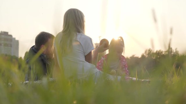 Happy caucasian family sitting on the meadow in sunrays and eating apples. Father spending time with his teen daughter and little blonde girl outdoors.