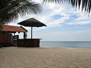 Wooden bar counter with an umbrella made of natural materials. On snow-white beach sand against the background of the sea. Hanging palm trees create comfort in a special way