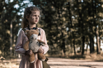 cute kid holding dirty teddy bear near trees in chernobyl, post apocalyptic concept
