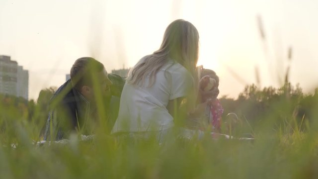 Two caucasian girls and their father sitting on the meadow in sunrays and eating apples. Happy family spending time together outdoors.