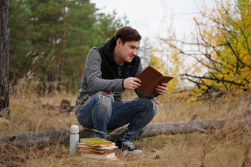 Fototapeta premium A young handsome man in casual clothes is sitting on a log and reading a book in the autumn forest.