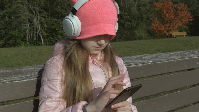 Pretty Cute Little Girl in Headphones and with smartphone on bench